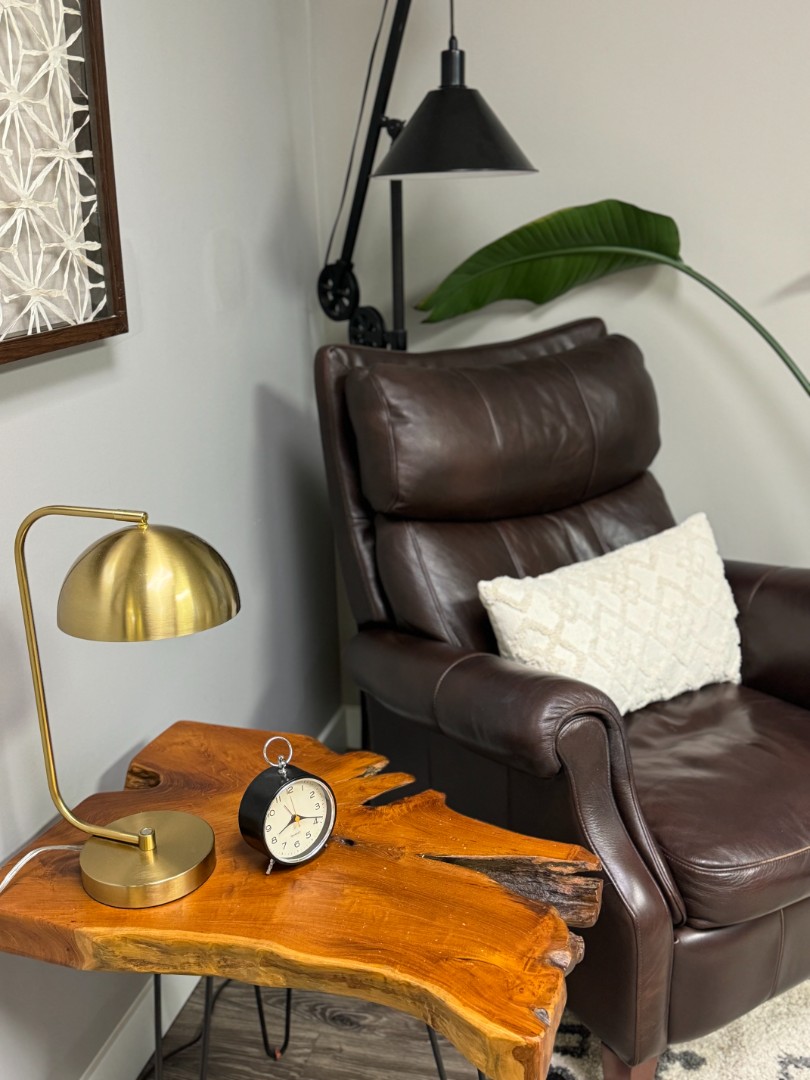 A brown leather chair with a lamp hanging over it. An end table made of a piece of tree with a lamp and clock on it.